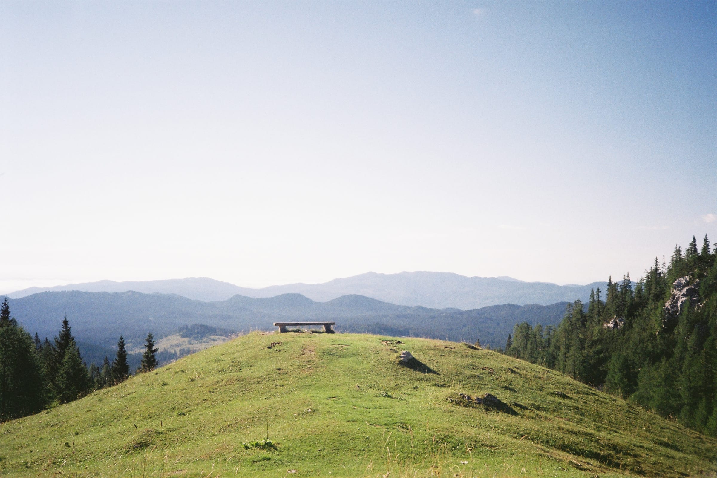 A grassy summit with a wooden bench, forested ridges fading into haze beyond